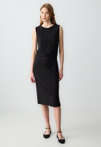Schwarzes ärmelloses Kleid mit vertikalen goldenen Streifen, knielang, tailliert. Glattes Material mit rundem Ausschnitt. Kombiniert mit schwarzen Ballerinas.