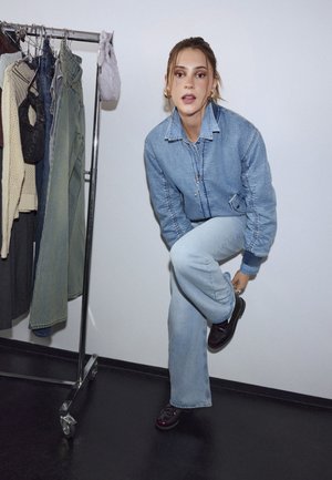 Nő farmerdzsekiben és farmerral cipőjét kötözi, miközben egy ruhaszárító mellett áll, amelyen nadrágok és pulóverek lógnak.