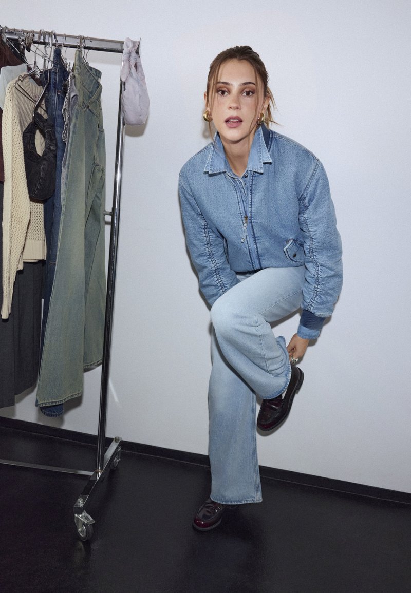 Moteris, dėvinti džinsinę striukę ir džinsus, užsisega batą stovėdama šalia drabužių stovo su kabančiomis kelnių ir megztinių.