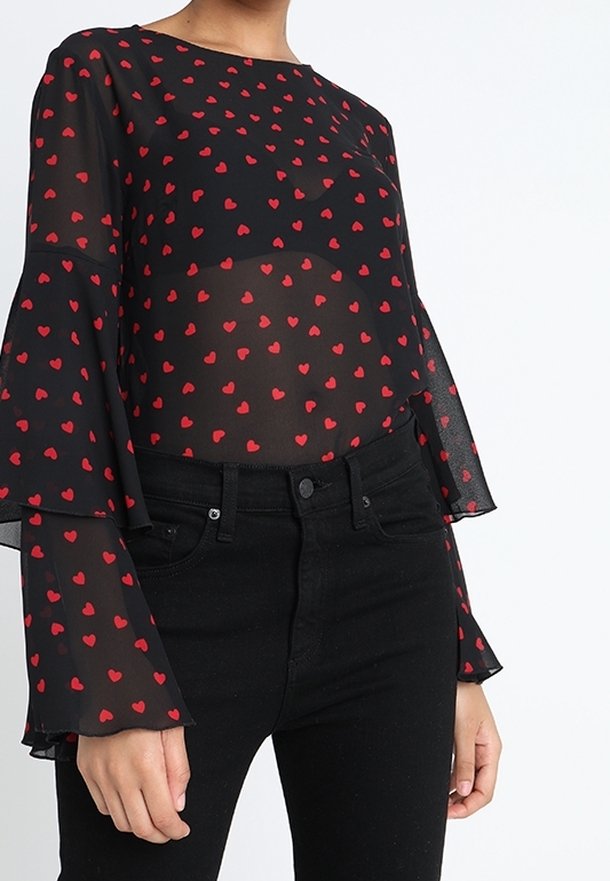 Blouse noire transparente avec manches longues évasées, ornée d'un motif dispersé de petits cœurs rouges. Associée à un jean noir taille haute.