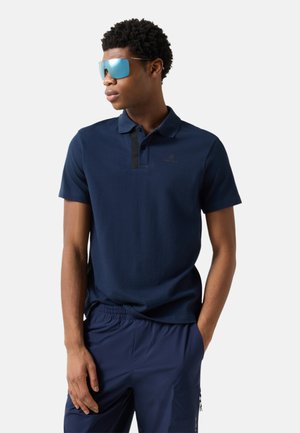 Młody mężczyzna w ciemnoniebieskiej koszulce polo, dopasowanych spodniach i refleksyjnych niebieskich okularach przeciwsłonecznych, stoi z jedną ręką w kieszeni.