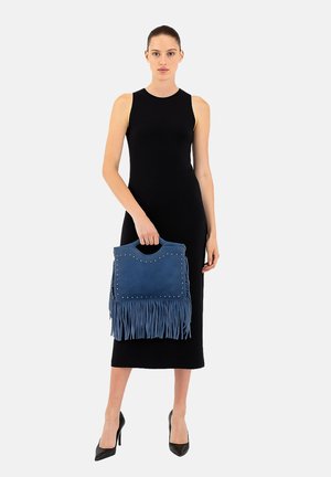 Een vrouw in een mouwloze zwarte midi-jurk en zwarte hakken, die een blauwe handtas met franjes en metalen studs vasthoudt voor een egale achtergrond.