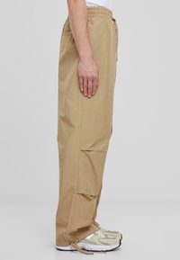 Beige, lichtgewicht, stoffen broek met een elastische tailleband, zijzakken en een losse pasvorm. Gecombineerd met grijze en witte sport schoenen.