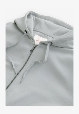 Svijetlosivi zip-up dukserica s kapuljačom na vezanje, glatke teksture i srebrnim dodacima. Ima oznaku na unutarnjoj ogrlici.