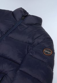 Mørkeblå puffjakke med høj krave, lynlås-lukning og et rundt logo-tryk på ærmet. Har quiltet tekstur og glat overflade.