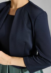 Chaqueta corta de color azul marino con una textura suave, hombros estructurados y mangas de tres cuartos, combinada con el escote de un vestido a juego.