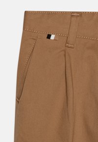 Khaki byxor gjorda av texturerad bomullstwill, med veck, bred midja och en liten svart och vit etikett nära toppen.
