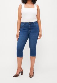 Capri in denim di colore blu scuro con design aderente, vita media e due tasche frontali. Abbinati a un top bianco con scollatura a quadrato e tacchi con stampa animale.