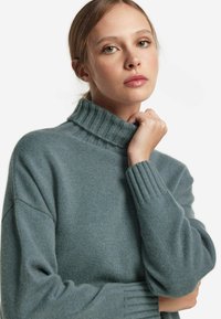 Mjukt tealfärgat polotröja med ribbad halsringning och muddar. Tyget verkar texturerat och lätt, lämpligt för lager-på-lager.