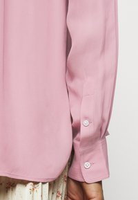 Långärmad blus i mjuk rosa, med en slät textur, knappärmar med vita knappar och rundad fåll.