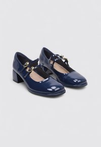 Zapatos Mary Jane de charol azul marino con tacones de bloque y correas de doble hebilla dorada, posicionados en ángulo uno al lado del otro sobre un fondo gris claro.