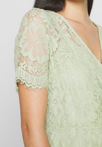 Robe en dentelle vert clair avec des motifs floraux, manches courtes à bord festonné et décolleté en V. Détails complexes sur l'ensemble.