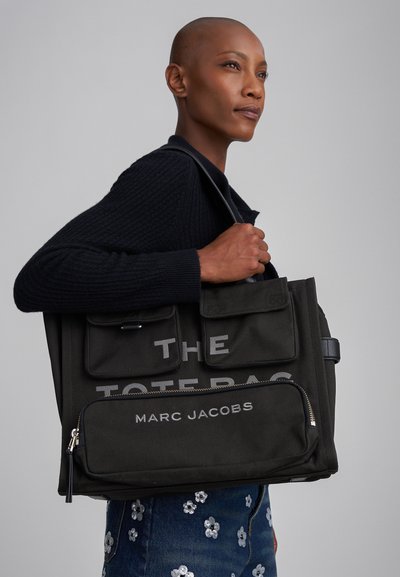 Donna con testa rasata, vestita con un maglione nero e jeans floreali, che tiene una grande borsa a tracolla nera di Marc Jacobs.