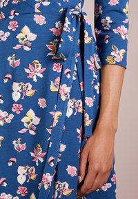 Blå klänning i omslag med blommönster i rosa, gult och vitt, med en knytning vid midjan och trekvartslånga ärmar.