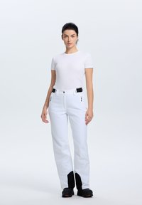 Pantaloni da sci bianchi con un design slim, tasche frontali e dettagli neri ai polsini. Abbinati a una maglietta bianca aderente e stivali da sci neri.