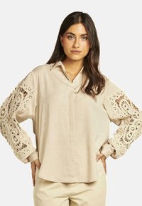 Blusa beige con cuello acampanado y mangas largas que presenta intrincados recortes de encaje. Corte ligeramente holgado con un dobladillo curvado.