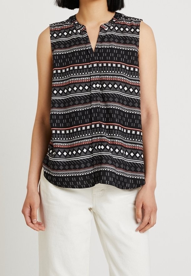 Haut noir sans manches avec des rayures géométriques rouges, blanches et grises, porté avec un pantalon blanc par une personne aux cheveux noirs courts.
