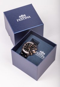 Montre noire avec des accents en or rose, boîtier en acier inoxydable et fonctionnalités de chronographe, présentée dans une boîte bleue avec un logo blanc.