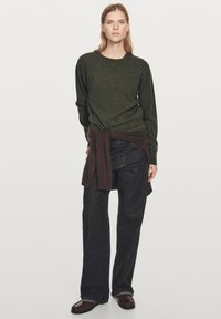 Femme debout portant un pull vert foncé, un jean noir à jambes larges, un pull marron noué autour de la taille et des mocassins marron.