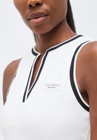 Κοντινό πλάνο ενός ατόμου που φορά λευκό αθλητικό αμάνικο τοπ με μαύρο περίγραμμα και λογότυπο "Ted Baker Sport" στο στήθος.