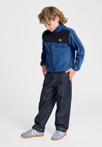 Blauwe fleece jas met rits en zwart bovenstuk; gecombineerd met donkere denim jeans en lichtblauwe sneakers met witte accenten.