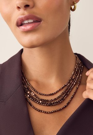 Mehrschichtige Halskette mit dunklen Perlen in verschiedenen Größen und facettiertem Design, getragen über einem dunklen Blazer.