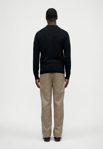 Homem com uma camisola preta de mangas compridas, calças texturizadas bege e sapatos pretos, de costas para a câmara, em um fundo simples.