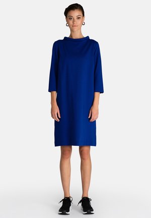 Blaues, knielanges Kleid mit Dreiviertelärmeln und Kapuze. Weicher Stoff, entspannte Passform. Kombiniert mit schwarzen Sneakers.