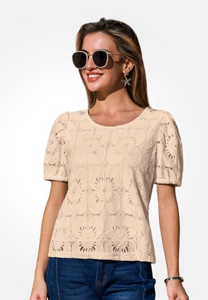 Vrouw met een beige kanten top met korte mouwen, donkere zonnebril, bloemige oorbellen, blauwe spijkerbroek en een armband, licht glimlachend naar de zijkant.