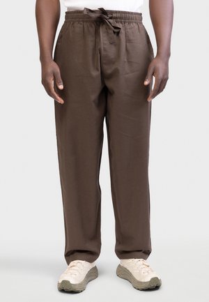 Άνδρας που φορά χαλαρά καφέ παντελόνια με κορδόνι και λευκά αθλητικά παπούτσια, στέκεται μπροστά σε απλό φωτεινό φόντο.