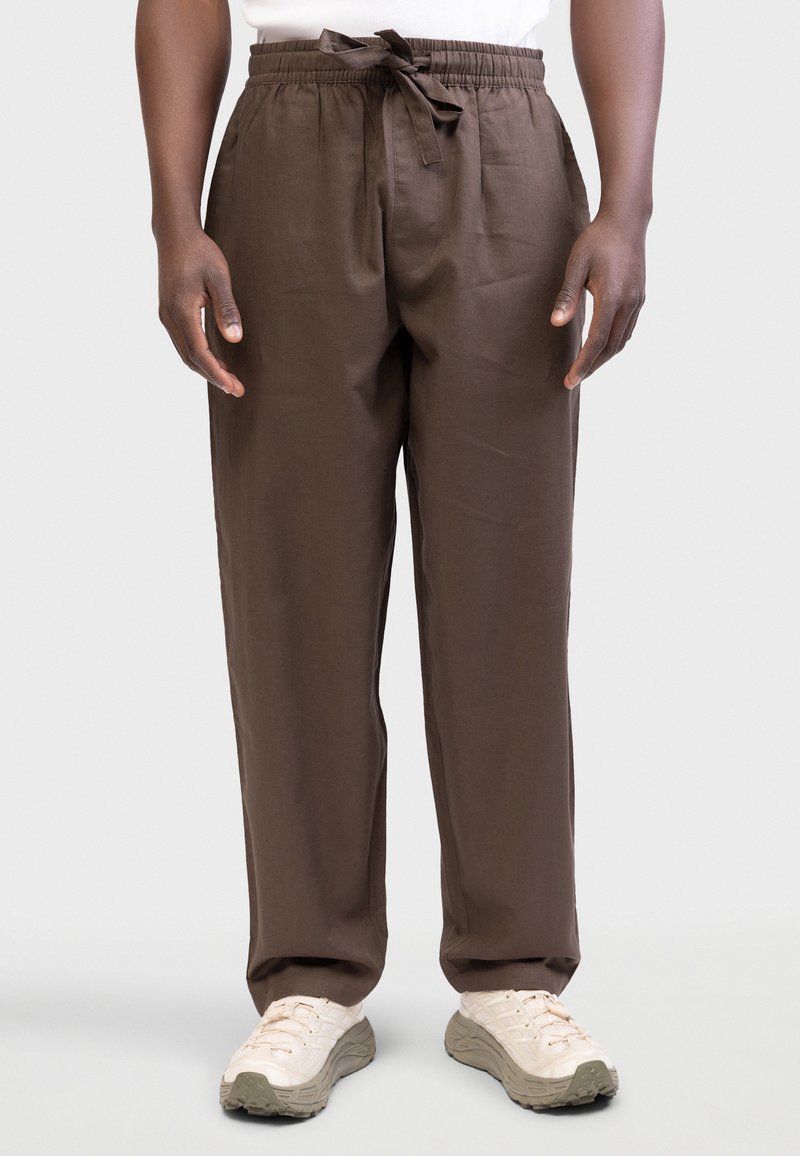 Άνδρας που φορά χαλαρά καφέ παντελόνια με κορδόνι και λευκά αθλητικά παπούτσια, στέκεται μπροστά σε απλό φωτεινό φόντο.