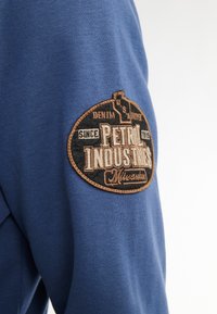 Felpa in cotone blu con un ricamo nero e marrone raffigurante la scritta "Petrol Industries" e "Milwaukee" sulla manica sinistra.