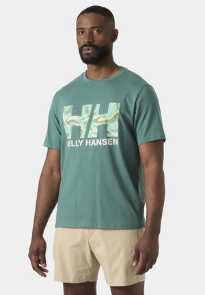 Hombre vistiendo una camiseta teal de Helly Hansen con un logo estampado y pantalones cortos beige, de pie contra un fondo claro y liso.
