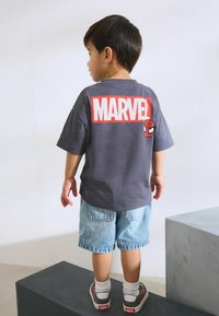 Szürke pamut póló piros "MARVEL" felirattal és Pókember grafikával a hátsó oldalán, farmer sorttal és színes sportcipőkkel párosítva.