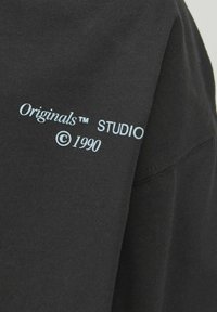 Svart bomullströja med ljusblå text broderad på ärmen, som lyder "Originals™ STUDIO ©1990." Enkel, avslappnad design.