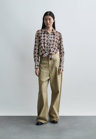 Γυναίκα που στέκεται δίπλα σε ανοιχτό γκρι τοίχο, φορώντας καφέ πουκάμισο με λουλούδια και κουμπιά, δεμένο στη μέση, και φαρδιά μπεζ παντελόνια με μικρό πουά μοτίβο.
