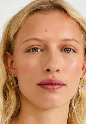 Boucles d'oreilles créoles colorées avec de petites perles vertes, bleues, roses et jaunes, sur un fond de peau claire.