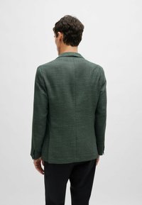 Blazer verde texturizzato con una vestibilità su misura, rever a punta e spacco posteriore singolo. Presenta polsini abbottonati e un design pulito e minimalista.