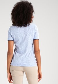 Ljusblå polo t-shirt med små röda prickar, kort ärm, och kontrasterande marinblå krage, visad från baksidan på en person som bär beige byxor.