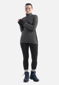 Giacca in pile zip-up nera e grigia con texture a coste, abbinata a leggins neri e scarponi da trekking blu con accenti grigi.