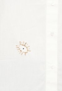 Λευκό πουκάμισο με μικρό κεντημένο σχέδιο ματιού από χρυσές και λευκές παγιέτες κοντά στα κουμπιά πάνω σε απλό ύφασμα.