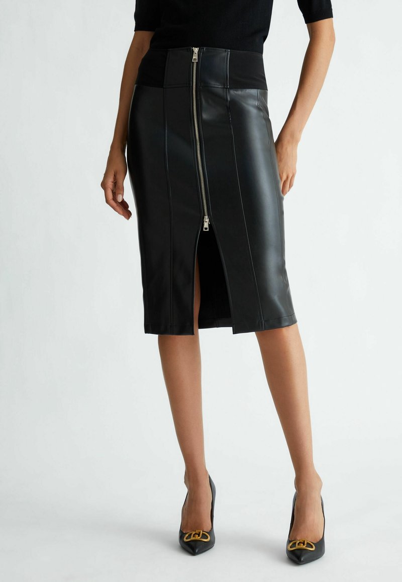 Femme portant une jupe en cuir noire longueur genou avec une fermeture éclair à l'avant et des talons pointus noirs avec des détails dorés, debout sur fond blanc.