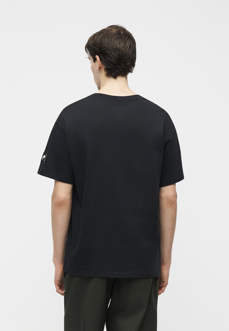 Junger Mann mit kurzen braunen Haaren, der ein schlichtes schwarzes T-Shirt mit kurzen Ärmeln und dunkelgrüne Hose trägt, steht mit dem Rücken zu weißem Hintergrund.