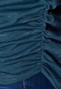 Haut en tissu bleu sarcelle avec une texture froncée, présentant des stries discrètes. L'ourlet passe à un jean en denim foncé.