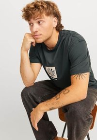 Camiseta de algodón verde oscuro con un diseño floral blanco en la parte frontal, combinada con jeans gris oscuro. Sujeto sentado en una silla de madera, apoyando la barbilla en la mano.