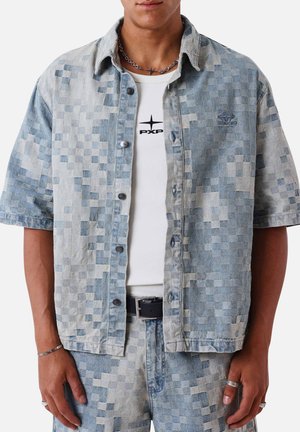 Mand iført blågrå pixelated kortærmet denim skjorte over hvid PXP-logo t-shirt med matchende pixelated denim bukser og sølvtilbehør.