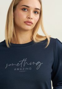 Maglietta a maniche lunghe blu navy con la scritta "QUALCOSA DI STUPENDO FREDDY" in testo argentato metallico. Tessuto morbido con scollo rotondo e vestibilità comoda.