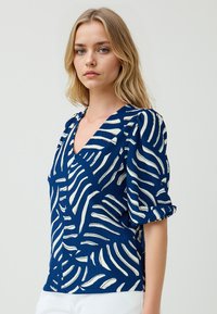 Blusa pattern navy con disegni bianchi a onde, dotata di scollatura a V e maniche corte a sbuffo. Tessuto testurizzato e design aderente.