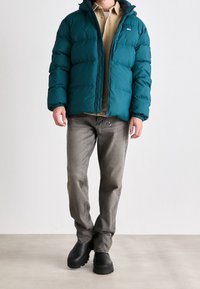 Teal pufferväst med huva, dragkedjestängning och elastiska ärmslut, som används över en beige skjorta, tillsammans med grå jeans och svarta stövlar.