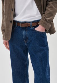 Jeans de mezclilla azul oscuro con un diseño de corte recto, con costuras claras y bolsillos frontales, usados con un cinturón de cuero marrón y una chaqueta de pana.